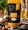 A stand-up pouch of Spice Professors Black Garlic Sea Salt is displayed on a rustic wooden surface surrounded by garlic bulbs, a bowl of coarse salt, and cooking ingredients in a warm kitchen setting.