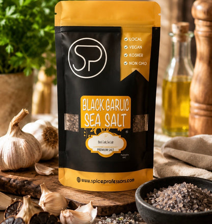 A stand-up pouch of Spice Professors Black Garlic Sea Salt is displayed on a rustic wooden surface surrounded by garlic bulbs, a bowl of coarse salt, and cooking ingredients in a warm kitchen setting.