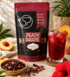 A stand-up pouch of Spice Professionals Peach Paradise hibiscus tea is shown on a tabletop beside a glass of red iced tea, with peaches, a bowl of loose tea, and tropical flowers in a styled setting.